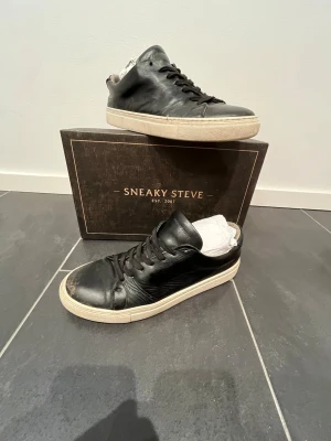 Sneaky Steve svarta sneakers i skinn - Sneaky Steve Moore Leather Shoe i svart skinn, storlek 42. Klassiska sneakers med rund tå, snörning och vit platt sula. Snygg minimalistisk design som funkar till många stilar. Tillverkade i Portugal och kommer med originalkartong.