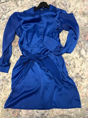 Blå satin look klänning med rosett - Snygg blå kortklänning från Bikbok med långärmade ärmar, knappar framtill och en stor rosett i midjan. Klänningen har klassisk skjortkrage och en diskret dragkedja i sidan för enkel påklädning. 