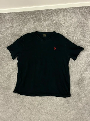 Svart t-shirt med röd logga från Polo Ralph Lauren - Svart t-shirt från Polo Ralph Lauren med klassisk röd broderad logga på bröstet. T-shirten har v-ringad hals och korta ärmar. Tillverkad i mjuk bomull som känns skön mot huden. Perfekt för dig som gillar stilrena och enkla plagg med en touch av lyx. Tröjan är använd så slitage finns 