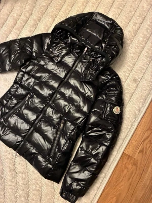 Svart Moncler pufferjacka med huva - Säljer en svart pufferjacka från Moncler med glansig finish och huva. Passar en XS och S, finns även QR kod som garanterar äktheten. 