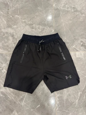 Svarta träningsshorts från Under Armour - Snygga svarta träningsshorts från Under Armour med elastisk midja och snörning. Shortsen har dragkedjeförsedda fickor, ventilerande hål på sidorna och logga framtill. Perfekta för gymmet eller löpning, tillverkade i lätt polyester för maximal komfort.