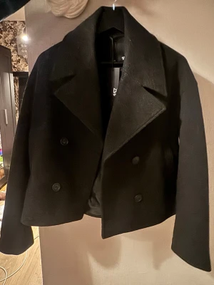 Svart kappa från 157 XS - Säljer en svart kappa från 157 i storlek XS. Jackan har dubbelknäppning med stora knappar, bred krage och en croppad, loose passform. Materialet är ull och ger en lyxig känsla. Perfekt för dig som gillar stilrena och klassiska plagg. Aldrig använd, inköpt för 400kr
