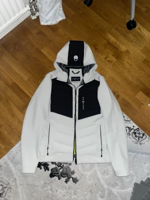 Sail Racing jacka - Säljer en snygg pufferjacka från Sail Racing i vit och svart med coola gula detaljer på insidan. Jackan har huva, dragkedjor och två stora svarta paneler framtill. Perfekt för dig som gillar sportig och modern stil.