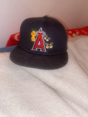 Svart New Era 59FIFTY LA Angels keps - Säljer en svart LA Angels 59FIFTY keps från New Era med broderade detaljer som blommor och fjärilar. Framsidan har ett stort rött A med halo, och sidan har World Series 2002 patch. Kepsen är tillverkad i 100% bomull och har klassisk flat brim. Lite smutsig går och tvätta enkelt!