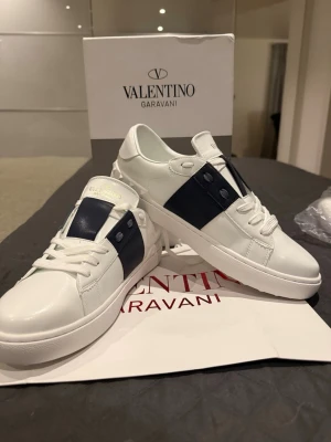 Valentino Garavani vita sneakers - Säljer ett par snygga Valentino Garavani sneakers i vitt skinn med marinblå detalj på sidan och coola nitar på hälen. Skorna har rund tå, platt sula och klassisk snörning. Perfekta för dig som gillar stilrena och lyxiga sneakers med en unik touch.
