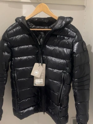 Svart pufferjacka från Canada Goose - Säljer en svart pufferjacka från Canada Goose med glansig finish och huva. Jackan har två stora dragkedjefickor framtill och märkets patch på ärmen. Perfekt för dig som vill ha en varm och snygg jacka med cool streetstil.