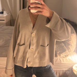 Beige kofta med stora knappar - Säljer en beige kofta med oversized passform och stora dekorativa knappar framtill. Koftan har två fickor och ribbade muddar vid ärmsluten. Perfekt för lager på lager och chill vardagsstil.