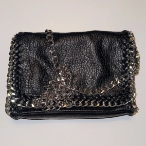 Svart axelväska med kedjedetaljer från Tiamo - Säljer en svart axelväska från Tiamo i skinnimitation med grov silverkedja runt kanterna och som axelrem. Väskan har dragkedja både på insidan och utsidan, samt snygga flätade detaljer längs kanten. Perfekt för dig som gillar en edgy stil.