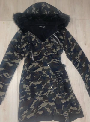 Camouflage jacka från Gothicana - Snygg parkajacka med camouflage-mönster i mörka toner från Gothicana. Jackan har en stor huva med svart fuskpäls, flera dragkedjor och knappar framtill samt ett bälte i midjan. Står som storlek XL men mer som en M/S🍭 använd fåtal gånger