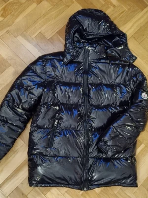 Svart Moncler pufferjacka  - Säljer en svart, glansig pufferjacka från Moncler med huva och dragkedja framtill. Jackan har quiltad design, två sidofickor med dragkedja. Inga tecken på slitage, bra skick.