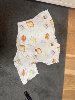 Vit t-shirt med frukostmönster från H&M - Rolig vit t-shirt från H&M med korta ärmar och tryck av frukostmotiv som ägg, toast och pannkakor. Rund hals med ljusrosa kant.  Det ingår även ett par matchande shorts till T-shirten! Perfekt för dig som gillar unika och lekfulla prints. Materialet är mjuk bomull som känns skönt mot huden.