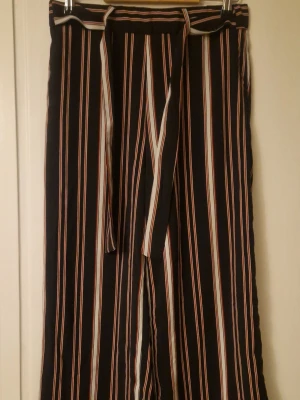 Randiga trekvartslånga byxor från Ellos - Snygga svarta wide byxor från Ellos i storlek 38 med vertikala ränder i vitt och rött. Byxorna har hög midja, elastisk linning och ett matchande knytband. Materialet är mjukt och lätt, perfekt för en chill och trendig look. Kolla gärna in mina andra annonser! 