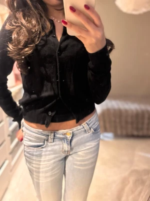 Svart kofta med knappar - Säljer en svart kofta med långa ärmar och knappar framtill. Tröjan är tunn och mjuk, perfekt att slänga över ett linne eller topp. Enkel och stilren design som passar till allt.