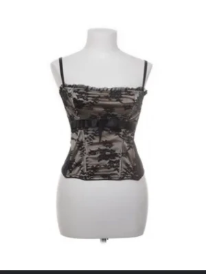 Svart och beige blommig korsett - Säljer en snygg korsett-topp i svart och beige med blommigt spetsmönster. Toppen har smala axelband, rosett framtill och genomskinlig spets över ett beige tyg. Perfekt att styla med jeans eller kjol för en cool look. St M= 40/42