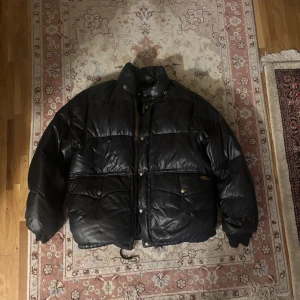 Vintage Svart pufferjacka från Polo Ralph Lauren - VINTAGE!!! Svart pufferjacka från Polo Ralph Lauren med hög krage, stora fickor med knappar och glansig finish. Jackan är tillverkad i polyester och har en klassisk, oversized passform som ger en cool och avslappnad vibe. Perfekt för kalla dagar när du vill hålla stilen.