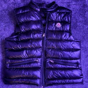 Svart Moncler Gui Väst - Svart Moncler Gui väst i storlek M, otroligt bra skick och väl omhändertagen. Original tags (bild 5/5) medföljer, nfc chip i loggan finns samt qr-sqan (bild 3/5). Hör av dig vid frågor och funderingar. Pris är ej hugget i sten 🤝