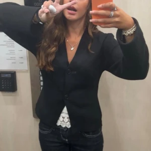 Svart cardigan jacka med knappar - Svart cardigan jacka från Ginatricot med v-ringad hals och två stora knappar framtill. Jackan har långärmade, lite utsvängda ärmar med knappdetalj vid mudden. Mjuk stickad känsla och clean, stilren look som är lätt att matcha med jeans eller kjol.