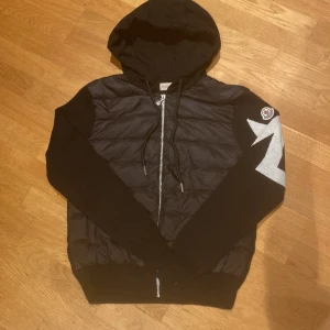 Svart pufferjacka Moncler med blixtdetalj - Svart pufferjacka från Moncler med huva och dragkedja framtill. Ena ärmen har en stor vit blixtdetalj och Moncler-logga. Jackan har quiltad front i blankt material och stickade ärmar och rygg. Snygg och unik design som sticker ut.