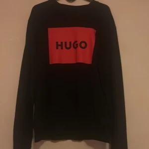 Svart långärmad tröja från Hugo(Boss) - Svart långärmad tröja från Hugo med en stor röd ruta och svart HUGO-logga på bröstet. Tröjan har en klassisk rund hals och är tillverkad i mjukt material som passar perfekt för chill dagar. Snygg och enkel design med fokus på loggan. Pris kan gå och diskuteras vid snabb och smidig affär!
