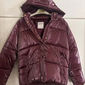 Vinröd pufferjacka - Säljer en vinröd pufferjacka från Minotti med glansig finish och huva. Jackan har guldiga knappar, dragkedja och två sidofickor. Perfekt för kalla dagar med sin tjocka, quiltade design. Storlek 152/158 vilket motsvarar Xs, men är rätt stor i storleken. Mycket bra skick, bara använd ett fåtal gånger 