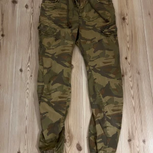 FSBN camouflage cargopants S - Säljer ett par snygga cargopants från FSBN i storlek S. Byxorna har ett klassiskt camouflage-mönster i gröna och bruna toner, flera fickor med knappstängning och snörning i midjan. Perfekt för dig som gillar streetwear och funktionella detaljer.