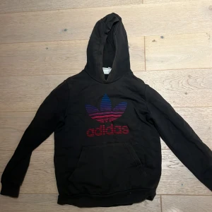 Svart Adidas hoodie med färgglad logga - Svart hoodie från Adidas med stor färgglad Trefoil-logga i blått, lila och rött på bröstet. Klassisk känguruficka framtill och huva. Tillverkad i mjuk bomull, perfekt för chill dagar eller streetstyle.