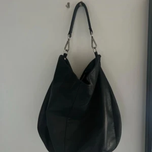 Svart axelväska i skinn med dragkedja - Stilren svart axelväska i mjukt skinn med silverfärgade metalldetaljer och praktisk dragkedja upptill. Väskan har en bred, avtagbar axelrem och rymlig form som gör den perfekt för att bära med sig allt man behöver. Enkel och snygg design som passar till många outfits.