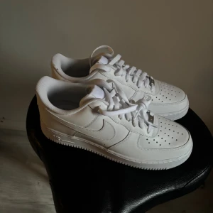 Nike Air Force 1 vita sneakers - Aldrig använda, Fräscha Nike Air Force 1 sneakers i helvitt skinn med klassisk låg siluett. Skorna har perforerad tå, snörning och platt sula. Snygg och tidlös design med Nike-logga på sidan. Perfekta för dig som gillar cleana och stilrena sneakers.