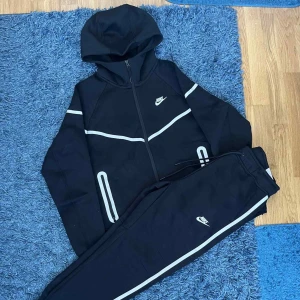Svart Nike reflective tech fleece dress  - Svart vindjacka från Nike med vit logga på bröstet och vita detaljer längs ärmar och fickor. Jackan har huva, hel dragkedja och sportig design. Materialet är polyester och passformen är normal. Perfekt för dig som gillar streetwear och sportig stil.