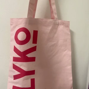 Rosa shoppingväska från Lyko - Snygg rosa tygväska från Lyko med coolt röd texttryck på båda sidor. Perfekt storlek för att bära böcker, laptop eller snacks. Väskan har två handtag och är gjord i slitstark bomull. Trycket 'Your beauty playground' ger den extra attityd.