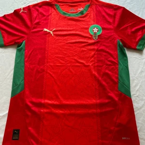 Marocko fotbollströja Puma röd/grön - Snygg Marocko landslagströja   i rött med gröna detaljer på krage och sidor. Kortärmad modell med guld Puma-logga och Marockos emblem på bröstet. Tillverkad i lätt polyester med subtila stjärnmönster i tyget. Perfekt för match eller häng med polarna.