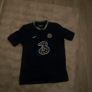 Chelsea FC marinblå fotbollströja Nike - Snygg marinblå Chelsea FC fotbollströja från Nike med klubbmärke på bröstet och stor vit sponsorlogga. Tröjan har korta ärmar och rund hals med mönstrad insida. Perfekt för dig som älskar fotboll och vill visa ditt stöd för Chelsea.