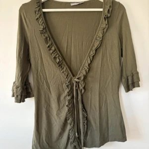 Olivgrön omlottopp med volang från Lindex - Säljer en olivgrön omlottopp från Lindex i storlek S. Toppen har volangkant längs ringningen och ärmarna, samt knyt framtill. Materialet är mjukt. Ärmarna är trekvarts och toppen har en smickrande v-ringad design.