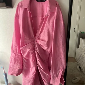 Rosa omlottklänning från Zara - Säljer en rosa kortklänning från Zara med snygg omlottdetalj och drapering framtill. Klänningen har lång ärm, klassisk krage och är tillverkad i ett lätt bomullsmaterial. Perfekt för dig som gillar färg och vill sticka ut lite extra.