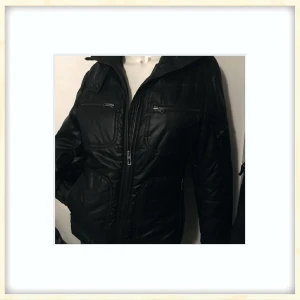 Svart pufferjacka från Marc O'Polo - Svart pufferjacka från Marc O'Polo med flera praktiska fickor med dragkedja och hög krage. Jackan är tillverkad i 100% polyester och har en snygg quiltad look. Perfekt för dig som vill ha en stilren och funktionell jacka med coola detaljer.