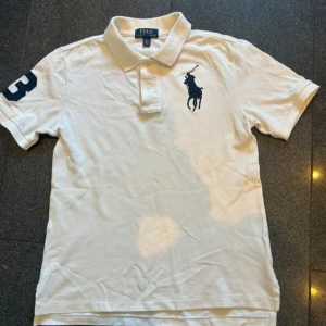 Vit pikétröja från Polo Ralph Lauren - Vit pikétröja från Polo Ralph Lauren med klassisk krage och knappar framtill. Stor mörkblå broderad logga på bröstet och en mörkblå siffra 3 på ena ärmen. Tröjan är gjord i bomull och har en normal passform.