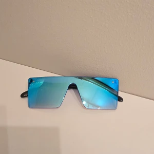 Blå spegelglas solglasögon - Säljer ett par futuristiska solglasögon med stora, rektangulära blå spegelglas och svarta skalmar. Glasögonen har en cool, kantig design som sticker ut och passar perfekt för dig som vill ha en unik stil.