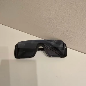 Svarta futuristiska solglasögon - Säljer ett par svarta solglasögon med stor, rektangulär sköldformad lins och markerad båge. Glasögonen har en modern och sportig vibe med mörka glas och små silvriga detaljer på sidorna. Perfekta för dig som vill sticka ut med en edgy stil.