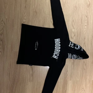 Svart HOODRICH hoodie med tryck - Svart hoodie från HOODRICH med stora vita tryck på bröstet och huvan. Klassisk känguruficka framtill och coola detaljer med text även på ryggen. Perfekt streetwear-vibe och skön passform med långa ärmar och huva.