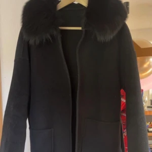 Kappa med ä*ta päls !  - Säljer en svart kappa med fluffig päls runt kragen och ett matchande bälte i midjan. Jackan har långa ärmar och stora fickor framtill. Perfekt för dig som vill ha en stilren och mysig look under höst och vinter.