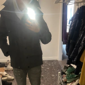 Grå Canada Goose Wyndham  - Säljer en grå dunjacka från Canada Goose med ikonisk röd och vit patch på ärmen. Jackan har huva, knappar och dragkedja framtill. Perfekt för kalla dagar med sin tjocka och quiltade design. Stilsäker look med klassisk passform och snygga detaljer.