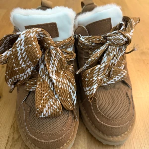 Bruna sneakers med fluffig plös - Säljer ett par bruna sneakers med breda, mönstrade snören i brunt och vitt. Skorna har en fluffig vit plös och detaljer i mocka och mesh. Sulan är platt och beige, och insidan ser riktigt mjuk ut. Perfekt för dig som gillar chunky stil och vill ha något som sticker ut.