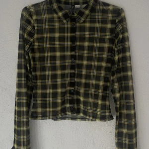 Rutig meshskjorta från H&M Divided - Snygg rutig meshskjorta från H&M Divided i svart och gul med transparent känsla. Skjortan har lång ärm, klassisk krage och knappar framtill. Croppad passform som ger en cool vibe. Materialet är lätt och stretchigt, 95% polyester och 5% elastan.