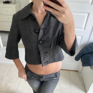 Grå croppad kavaj med mönster - Snygg grå croppad kavaj med fiskbensmönster och stora knappar framtill. Jackan har bred krage och trekvartsärmar som ger en cool och modern vibe. Perfekt att styla med jeans eller kjol för en trendig look.