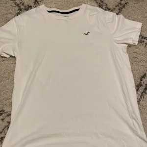 Vit t-shirt från Hollister i bomull - Vit t-shirt från Hollister med klassisk passform och liten broderad logga i vinrött på bröstet. Tillverkad i 100% bomull, perfekt för en enkel och stilren look. Rund halsringning och korta ärmar. PRIS KAN DISKUTERAS! 