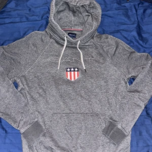 Grå hoodie från GANT med emblem - Säljer en grå hoodie från GANT i storlek L med broderat emblem och vita snören vid huvan. Tröjan har en klassisk känguruficka och ribbade muddar vid ärmslut och nederkant. Perfekt för chill dagar och snygg streetstil.