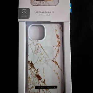 Carrara Gold Skal för iPhone 13 - Stilrent Carrara Gold skal till iPhone 13 från iDeal of Sweden. Marmormönster med gulddetaljer, MagSafe-kompatibelt. Skalet är nytt och levereras i originalförpackning, utan synligt slitage eller defekter. Perfekt för att skydda och ge din mobil en exklusiv look.