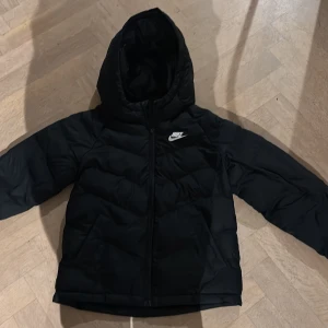Svart pufferjacka från Nike med huva - Svart pufferjacka från Nike med vit logga på bröstet. Jackan har huva, dragkedja framtill och quiltad design som ger en schysst look. Perfekt för kalla dagar och enkel att matcha med din stil.