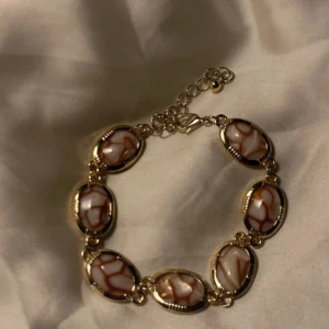 Guldigt armband med marmorerade länkar - Unikt armband med ovala länkar i guldton, dekorerade med marmorerade stenar i vitt och brunt. Länkarna har en snygg, detaljerad kant och armbandet har justerbar kedja för perfekt passform. Perfekt för dig som gillar statement-smycken med cool design.