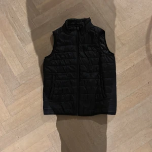 Svart dunväst från Everest - Svart quiltad dunväst från Everest med ståkrage och dragkedja framtill. Västen har två fickor på sidorna och en stilren, sportig look. Perfekt för lager-på-lager och passar dig som gillar enkel och funktionell stil. Den är lite tunnare i modell så den passar för lite varmare väder och sport.